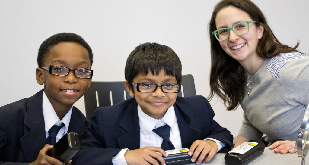 Two young boys in school uniforms sit and smile with their brand-new glasses, fitted by a woman eye doctor sitting to their left.