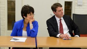 Two people, one woman and one man, sit listening to a presentation in a conference room. 