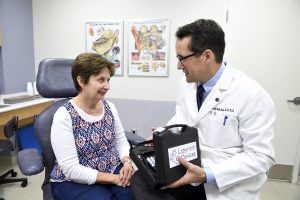 A female patient sits in a chair and speaks with a male doctor in a white lab coat, who holds a medium size black box that houses an electrical implant device.