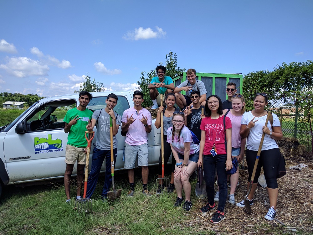 Twelve young men and women, some holding shovels and pitchforks, take a group photograph in front of a white truck parked in the middle of a farm field.