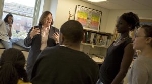 A woman speaks, gesturing with her hands and surrounded by a half-dozen high-school-aged students in a classroom setting.