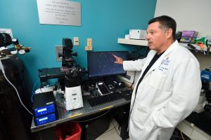 A man in a white lab coat stands in front of a microscope and a computer while pointing at an image on a monitor. 