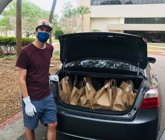 A man wearing a face mask and gloves stands behind a car that has its trunk open. Inside the trunk are several brown bags full of food to support health care workers at Johns Hopkins All Children's Hospital.