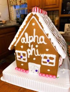 Brown gingerbread house with a peaked roof covered with white icing, red and white candy, and the words Alpha Phi