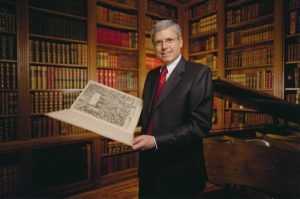 Bruce Fye, standing in front of a library that fills the walls, stands in front of the camera with a large open book with an ornate design on the page.