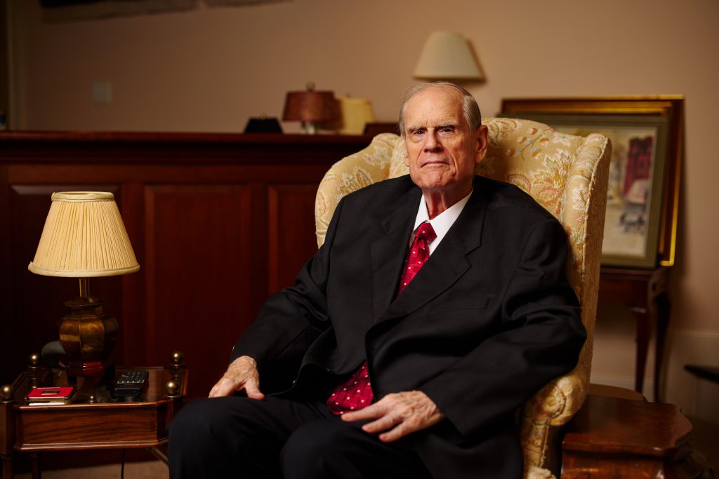 Wearing a dark suit, white shirt, and red tie, Wilmer Eye Institute and School of Medicine donor Philip Gerdine sits in a tan arm chair with dark paneling, a lamp, and a framed painting in the background.