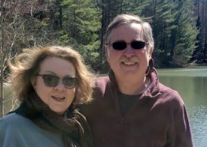 Linda and John stand side by side, wearing sun glasses and smiling in front of a lake.