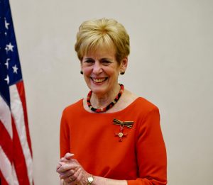 Standing by the U.S. flag, Jill McGovern smiles and wears a red dress with the Cross of the Order of Merit pinned to it.