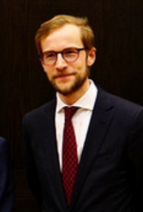 Former SAIS McGovern-Muller fellow Sebastian Ernst has a beard and wears glasses, white shirt, dark jacket and red tie.