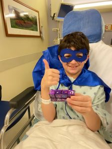 A young boy in a hospital bed wearing a superhero mask and cape holds a fidget toy from the fidget toy initiative in one hand and gives a thumbs up with the other.
