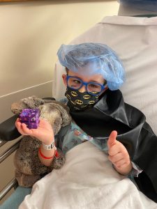 A young boy in a hospital bed wearing a face mask and cape holds a fidget toy from the fidget toy initiative in one hand and gives a thumbs up with the other.