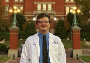 With the dome building in the background, Johns Hopkins medical student and HEAT Corps volunteer Andy Lancaster smiles in a blue shirt and white coat.