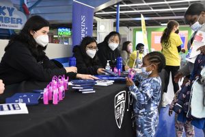 Three HEAT Corps volunteers in black sweatshirts and face masks sit at a table and interact with a preschooler in a mask and blue tie-dyed sweatsuit.