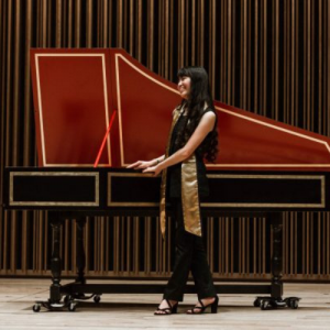 Standing in profile on a stage in front of a piano, Peabody Launch Grant recipient Mira-Fu-En Huang wears black pants, black top, and a long gold scarf.
