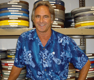 Geoff Alexander, wearing a blue Hawaiian shirt, smiles in front of a wall of archival films.