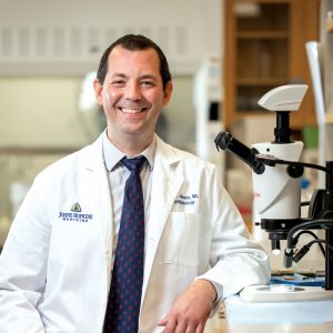 Wilmer Rising Professor Thomas Johnson wears a white lab coat over a light shirt and dark blue tie and smiles broadly as he stands next to optometry equipment.