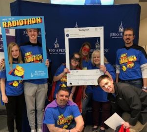 Wearing superhero capes and masks, eight Johns Hopkins Children’s Center Radiothon volunteers smile as they stand and hold Instagram and Radiothon cutout frames.