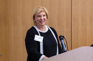 Wearing a black dress with white trim, School of Nursing Advisory Board chair Natalie Bush smiles and stands at a podium with a microphone.