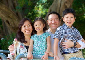 Jeffrey Shiu, Emily Liu, and their two children are standing and smiling in a park. Shiu is holding their son and Liu is standing behind their daughter.