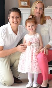 Wearing a pink and white dress, Laney Jaymes smiles and stands in between her parents Brian, wearing tan pants and a white shirt, and Laura Fitzsimons who wears pink pants and a white top.