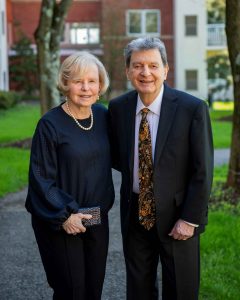 In front of a long driveway bordered by green grass and a tall tree and leading to a brick townhouse, Anne Ragonese smiles and wears blonde bobbed hair, a navy blouse and pearl earrings and necklace as she stands next to Paul Ragonese with short gray hair and wearing a navy blazer, white shirt and colorful tie.
