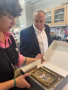 Michael Siegel who is wearing a white collared shirt and blue jacket is looking at preserved materials in the conservation lab. Jennifer Jarvis is standing to the left of Siegel showing him the items. 