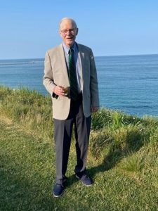 Rob Carr is standing outside smiling. There is a lake in the background. Rob is wearing a blue dress shirt, green tie, tan jacket, and dark dress pants.