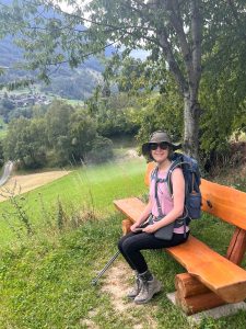 Amy Johnson is wearing hiking apparel with a blue backpack. She’s sitting on a wooden bench in a grassy area overlooking the Swiss Alps. 