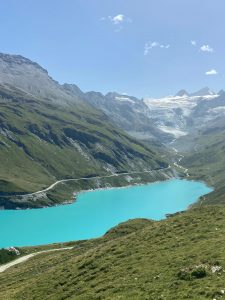 A lake sits below a range of the Swiss Alps. The mountains are snow-capped with a bight blue sky in the background.