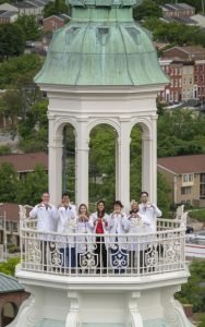 On top of the Johns Hopkins medicine dome, Cardiology Fellowship Program Director Thorsten Leucker stands to the left of six male and female fellows who wear white coats and hold their hands in the shape of hearts