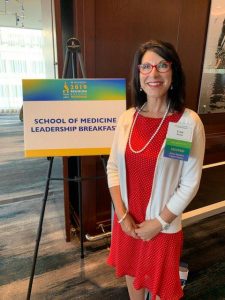 Lisa Bunin, MD, is standing, wearing red glasses, and smiling next to a School of Medicine Leadership Breakfast sign. She is wearing a red dress, white sweater with a pearl necklace.