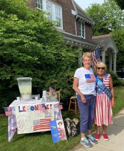 Suzette Warmus is wearing a patriotic dress with her Miss January sash. She is standing with Miss November. They are both smiling on the front lawn. To the left of them is the Fourth of July ALS lemonade fundraiser.