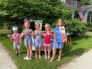 Suzette Warmus’s five grandchildren are standing outside on the lawn in front of their Fourth of July lemonade stand. They wearing patriotic outfits. The grandson is holding an American flag, while the granddaughters are holding an ALS sign, and Calendar Girls.