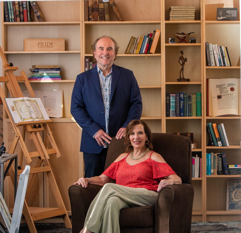 With airy, wooden bookshelves in the background, Randy Elrod stands and wears blue pants, blue jacket and blue and white printed shirt as Gina Elrod sits in a brown armchair wearing tan pants and a red blouse.