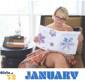The January calendar image. Suzette Warmus is wrapped in a colorful blanket, sitting on a light brown chair wearing Wendy's pearls. She's reading an oversized snowflake book. To the left is a nightstand with books stacked. It includes the Sheboygan North 1972 year book and ALS research book.