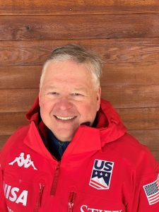 Peter Jennison is wearing a zipped up red ski jacket and smiling into the camera. He’s standing in front of a wood framed wall.