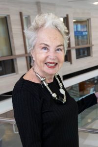 Monika Petter is standing and smiling in the lobby of an office. She's wearing a black dress with a silver necklace.