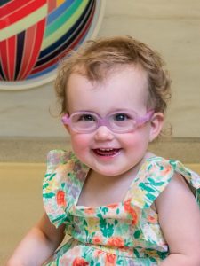 Johns Hopkins Wilmer Eye Institute pediatric vision patient Bernie Cowell Murray is one-year-old, has light brown curly hair and wears pink glasses and a sleeveless floral dress as she sits and smiles