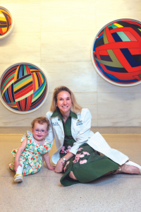 Sitting against a gray wall with circular colorful art in the background are one-year-old Wilmer pediatric vision patient Bernie Cowell Murray on left and Wilmer pediatric doctor Courtney Kraus