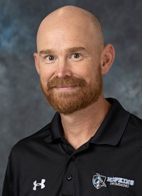 Scott Armstrong is smiling into the camera. He's wearing a black Hopkins swimming polo. He's sitting with a dark blue/gray backdrop behind him.