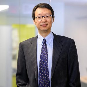 Pictured from the waist up, Carey Business School Senior Lecturer Xiaohua Hu stands wearing a dark navy pinstripe suit, light blue shirt, and navy tie