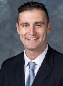 Dan Wodicka is smiling into the camera. He's sitting in front of a blue-gray backdrop wearing a dark jacket, with a white collared shirt, and blue tie.