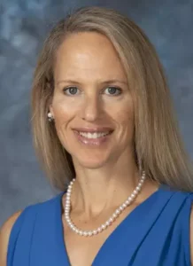 Jennifer Baker is smiling into the camera. She's sitting in front of a blue-gray backdrop and wearing a blue blouse with a pearl necklace.