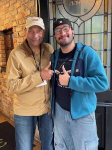 John Guess is standing to the left of Hadeed Habib. They are both giving the thumbs up sign. Guess is wearing a khaki baseball hat with a tan colored button down shirt with blue jeans. Habib is wearing a backwards black baseball hat with a black t-shirt, a blue fleece and jeans.