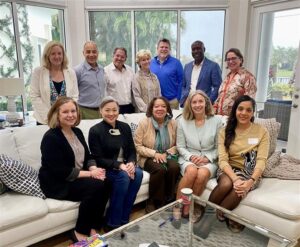 The School of Education National Advisory Council are gathered and smiling. Five of the members are sitting on a white couch while seven of the members are standing behind.