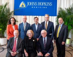 Underneath a blue Johns Hopkins Medicine sign, a woman in an orange jacket stands to the left of four men in suits and ties and above a man in a suit, a woman in a black pantsuit, and a man in a suit who are all seated