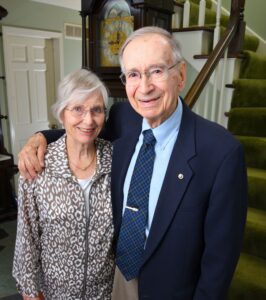 Karen and Clyde Flory, Jr., are standing and smiling inside their Michigan home. Karen is wearing a tan and white pattern blouse. Clyde is wearing a blue suit and has his arm around Karen.