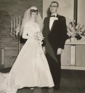 Karen and Clyde Flory, Jr., on their wedding day. Karen is wearing a white dress, vail, and holding a bouquet of flowers. Clyde is wearing a tuxedo. The couple is standing and smiling together inside a church.