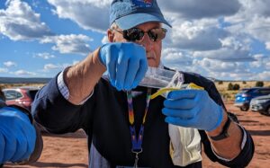 Against a desert background with a blue sky spotted with white clouds above it, Osprey Foundation president Bill Clarke wears blue gloves and pours a bottle of water into a glass