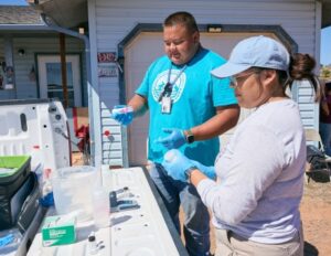 As Diné Navajo community members, a man wearing a bright blue shirt and women wearing a light blue hat stand outdoors next to a long table and test water samples